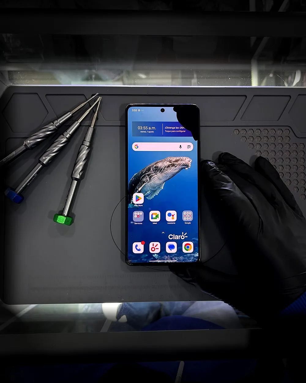 Mano con guante negro sosteniendo un celular con una zona de la pantalla sin imagen, sobre una alfombrilla de reparación y junto a tres destornilladores de precisión