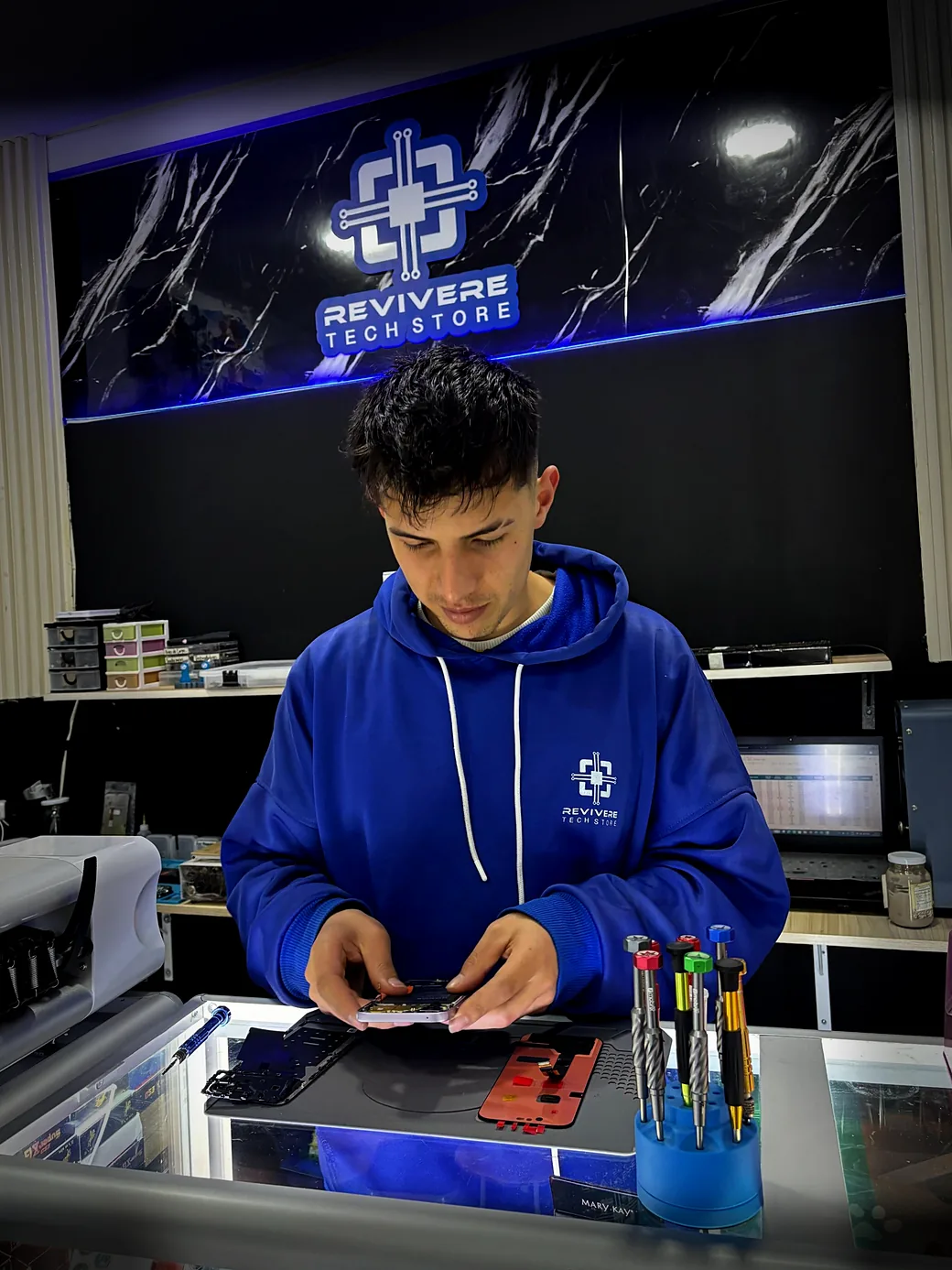 Técnico de Revivere revisando un celular en el banco de trabajo, frente al letrero de la tienda
