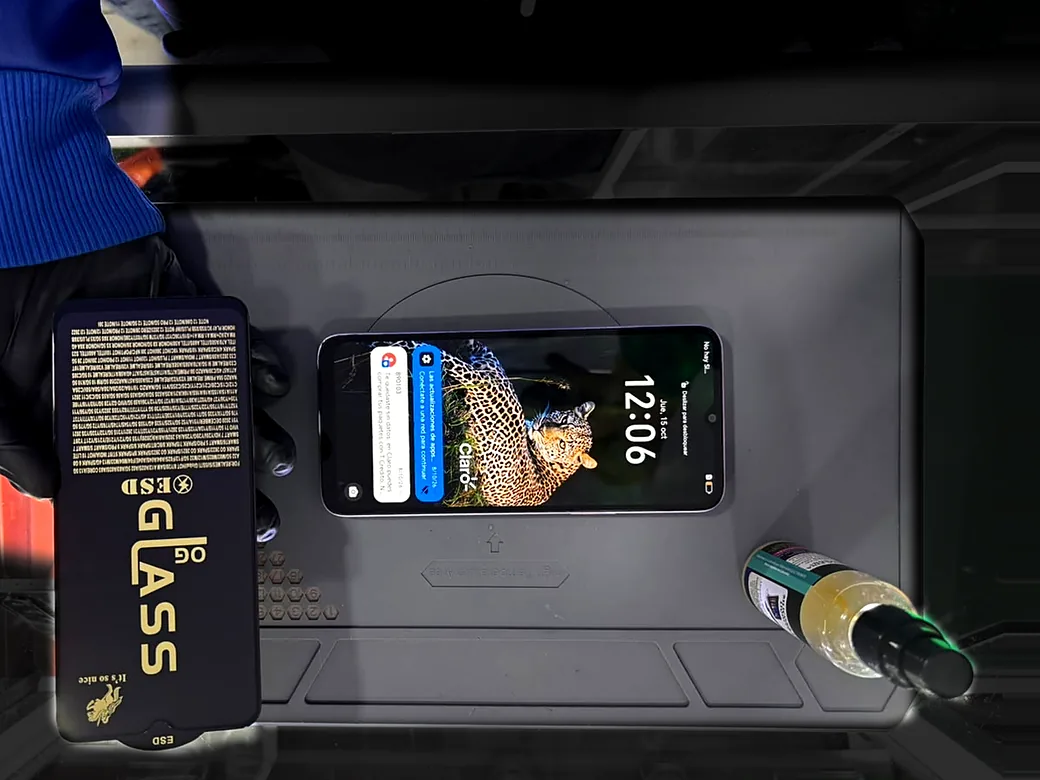 Mano con guante negro sosteniendo un vidrio templado junto a un celular con la pantalla encendida, sobre la alfombrilla de reparación